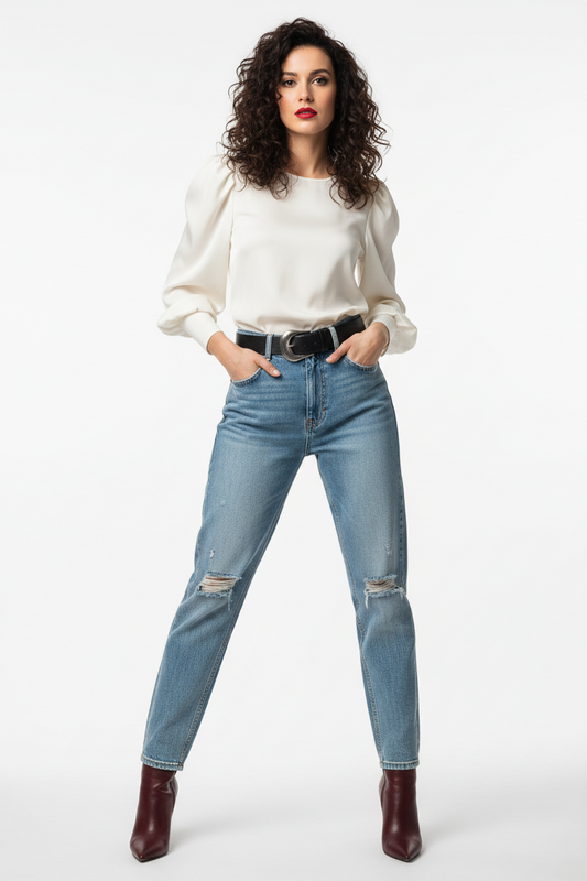 Vrouw in lichtblauwe high-waist jeans met scheuren op de knieën en zwarte riem, gecombineerd met een crèmekleurige satijnen blouse met pofmouwen en ronde hals, bordeauxrode laarzen, witte achtergrond.